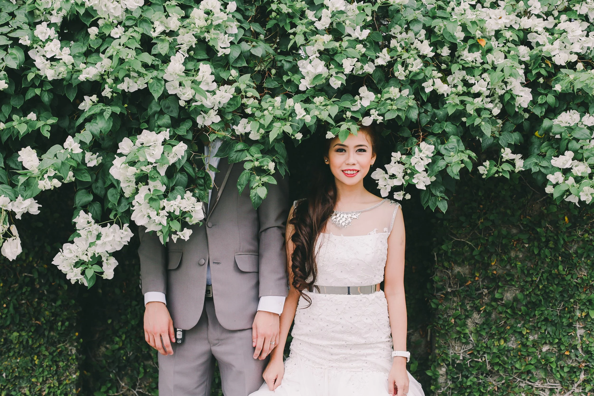 A woman in a white dress and a man in a gray suit are standing in front of a lush green bush with white flowers. The woman has long, dark hair and is smiling at the camera.