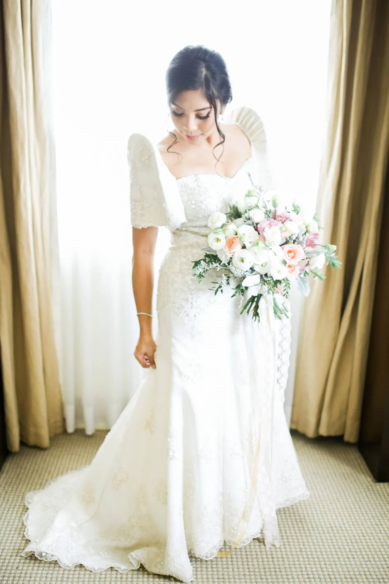 A bride is standing in front of a mirror, wearing a white wedding dress with lace details, holding a bouquet of white and pink flowers, with curtains framing the scene.