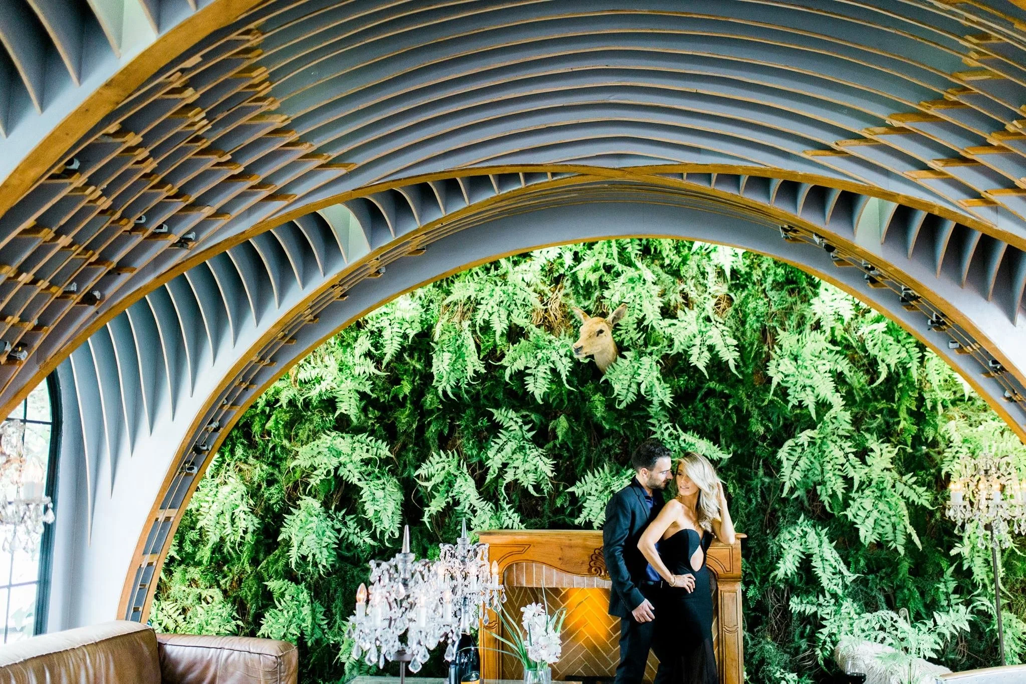 A couple in formal attire standing close together inside a green, plant-filled space with a curved architectural structure overhead.