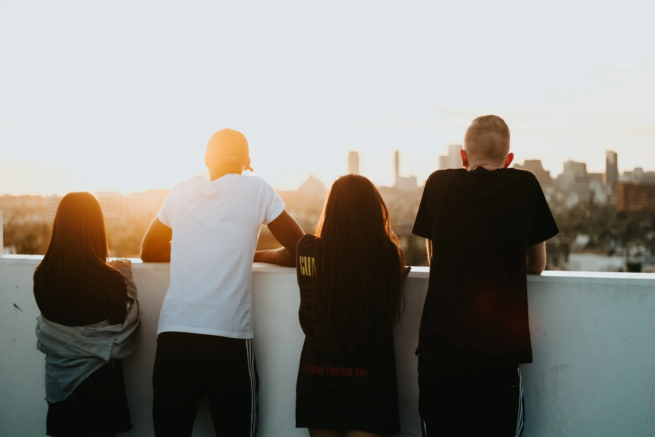 Four people stand on a rooftop overlooking a city skyline at sunset, facing away from the camera.