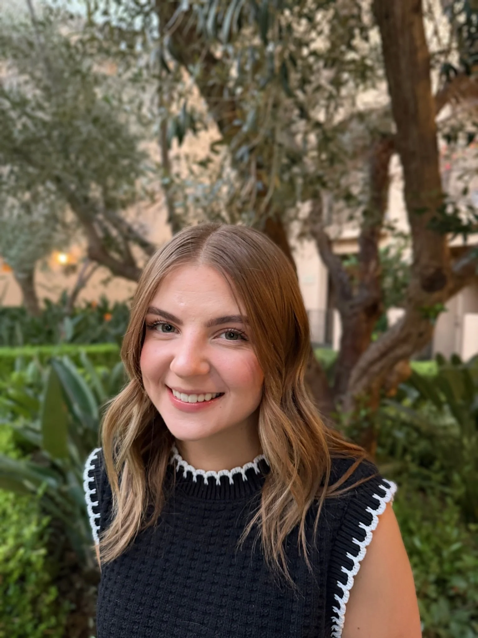 A young woman with long, wavy, light brown hair and light skin smiling outdoors. She is wearing a black top with white detailing around the neckline and sleeves. The background features a large tree with twisted branches and green foliage.