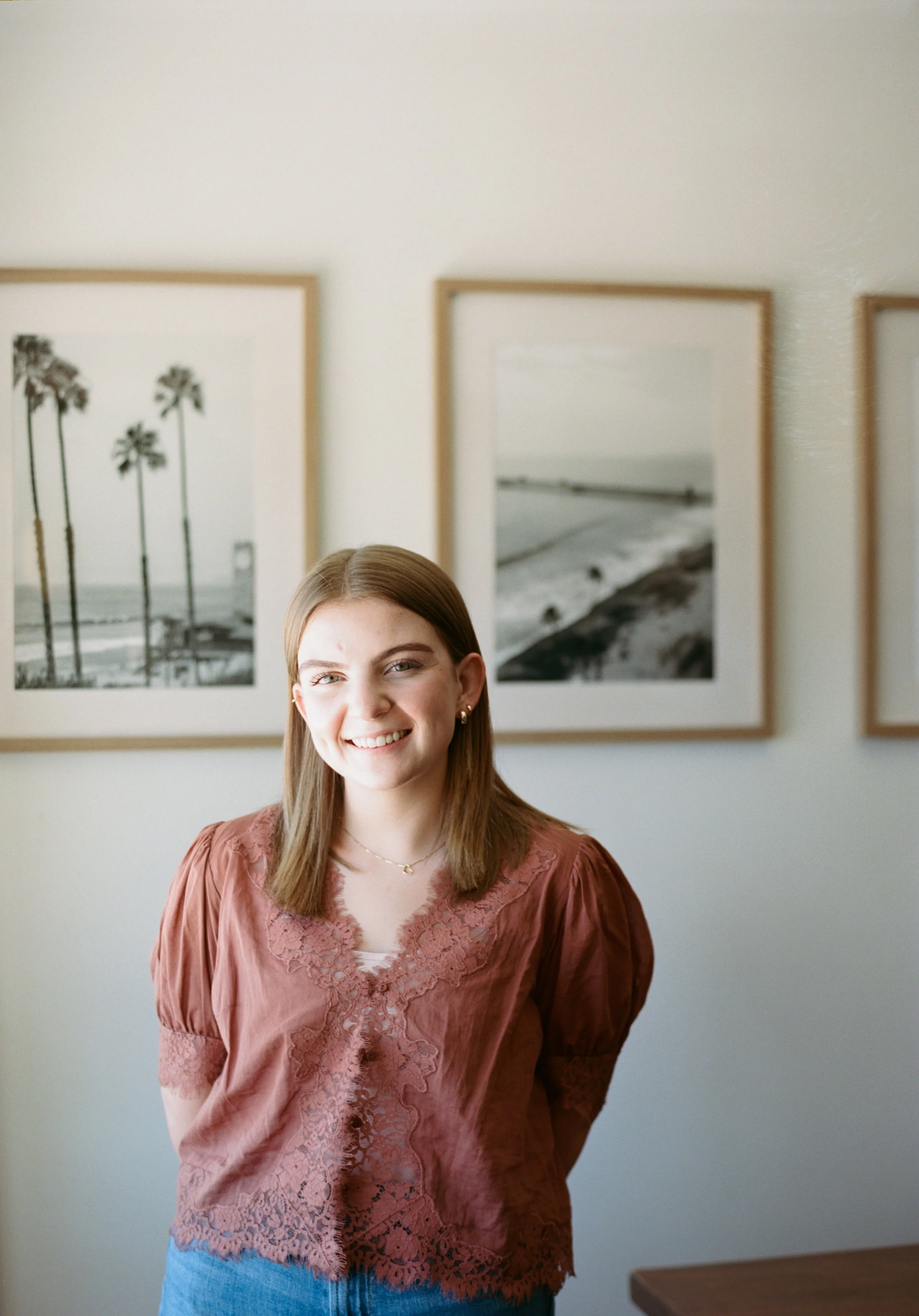 A young woman with shoulder-length light brown hair, smiling, standing in front of framed black and white photographs of beaches and palm trees on a white wall, wearing a salmon pink lace blouse and blue jeans.