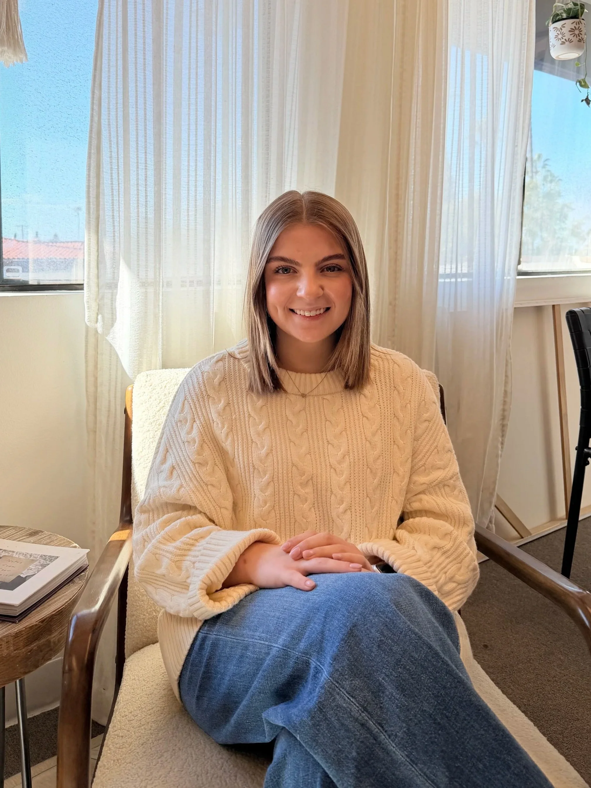 A young woman with shoulder-length blonde hair, wearing a beige knitted sweater, is sitting on a cream-colored chair in a room with light-colored curtains and large windows. She is smiling, with her hands resting on her lap, and looking at the camera.