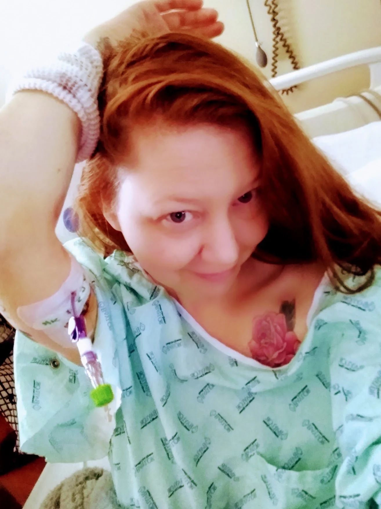 A woman lying in a hospital bed, with an IV inserted into her arm, wearing a hospital gown, and smiling at the camera.