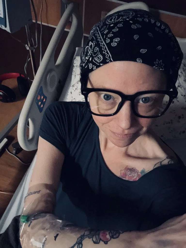A woman in a hospital bed with medical tubing attached to her arm, wearing glasses, a black bandana, and a black shirt, taking a selfie.