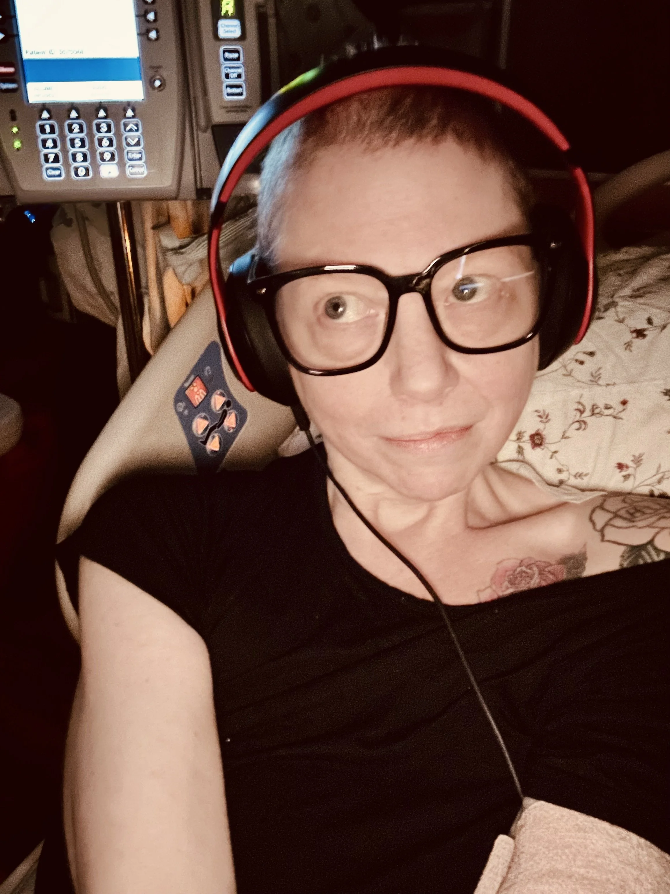A person with short hair, glasses, and tattoos, lying on a chair with a medical or spa device around their head and a control panel in the background.