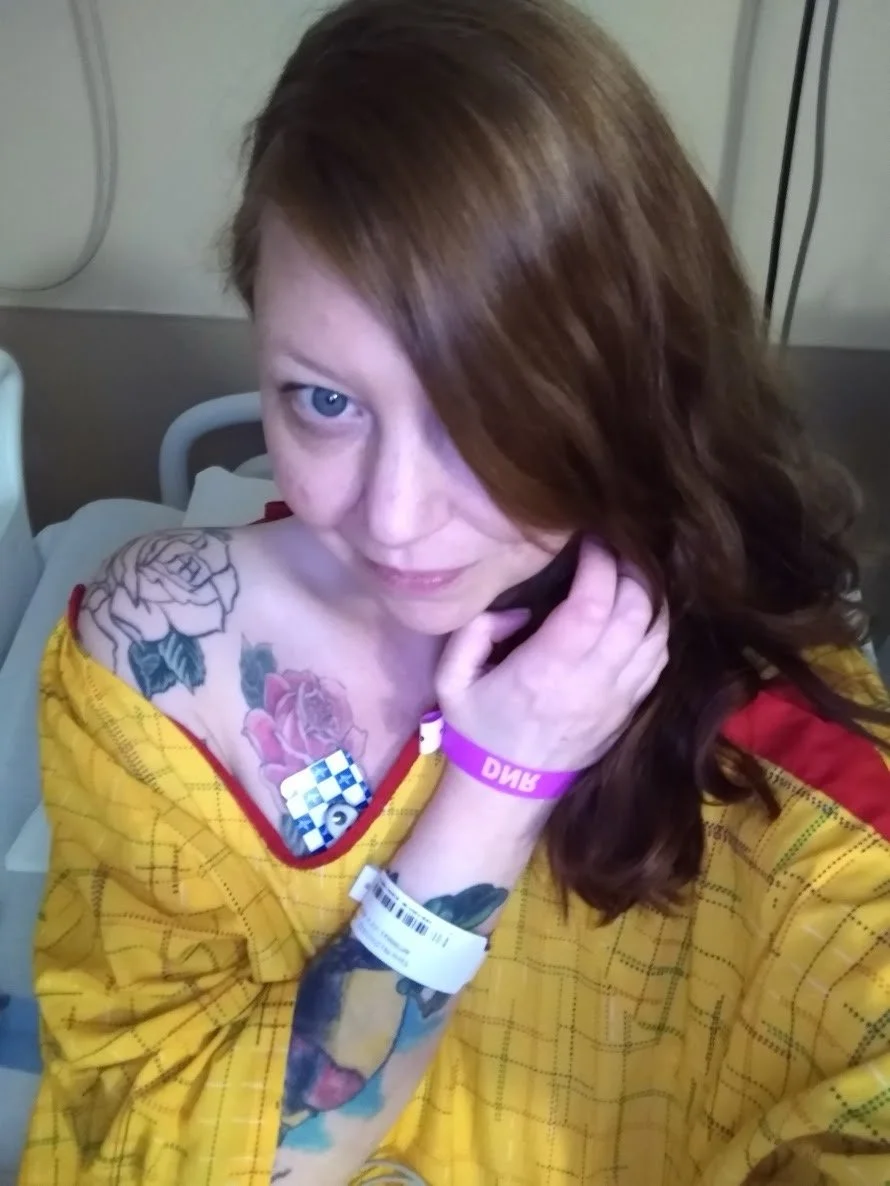A woman with red hair in a hospital gown, sitting in a hospital bed, showing tattoos on her shoulder and arm, with a hospital wristband and a card with a police badge logo.