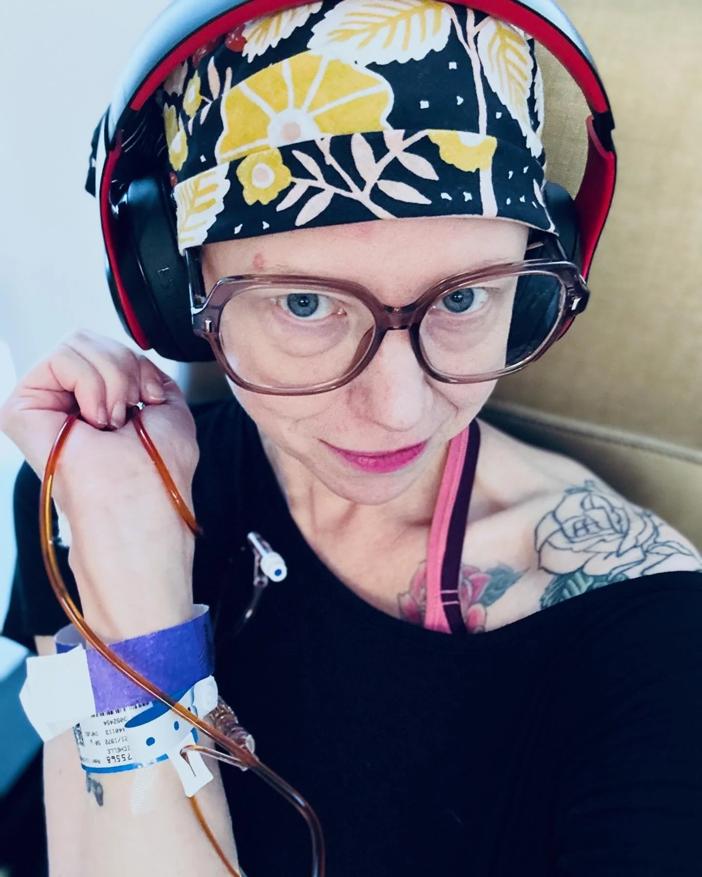 A woman with glasses and a floral headscarf looks at the camera while holding an IV insertion in her arm. She has tattoos on her shoulder and is in a medical setting.