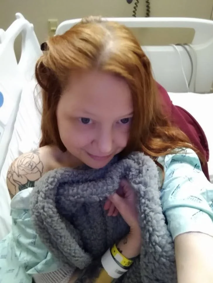 Young woman with red hair in a hospital bed, holding a grey fleece blanket, with hospital wristbands on her wrist.