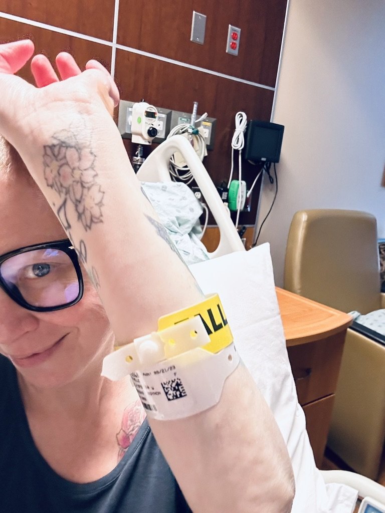 Woman in a hospital room with an IV bracelet on her arm, resting on a bed, wearing glasses and smiling.