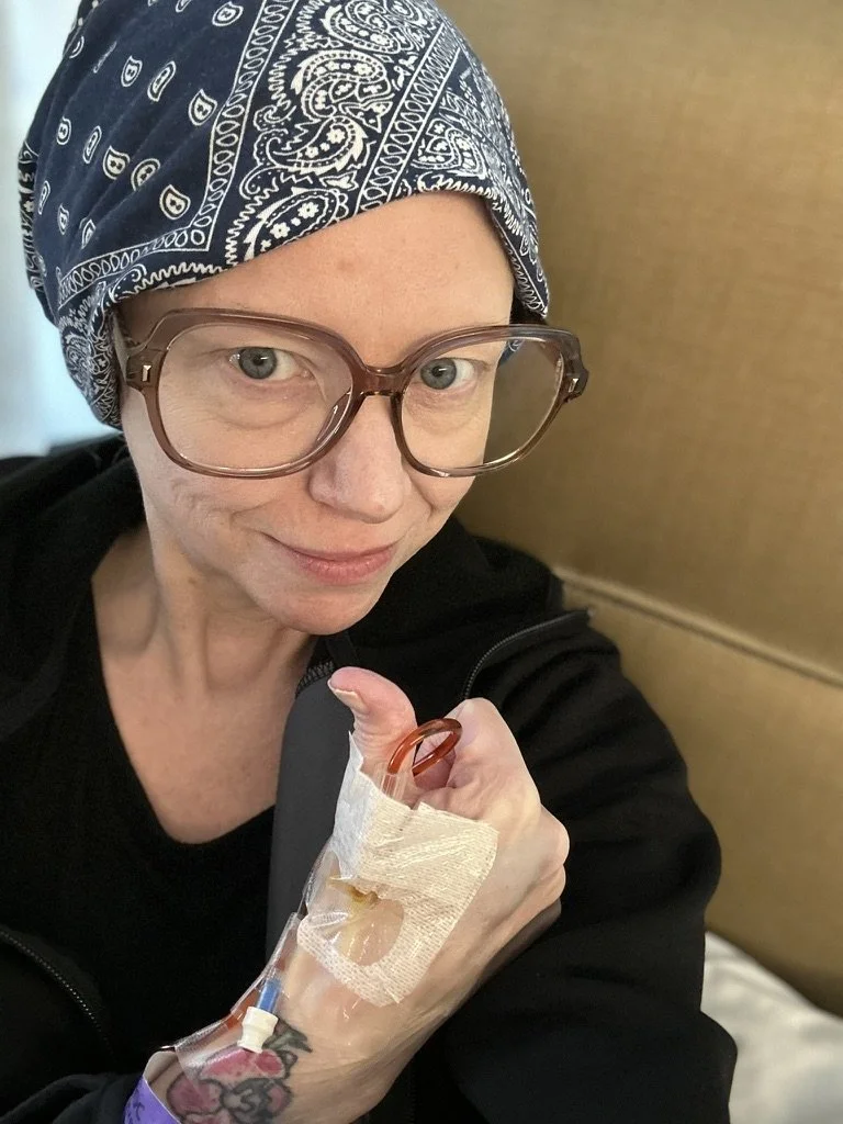 A woman wearing glasses and a blue bandana headscarf taking a selfie in a medical setting. She has a IV line in her hand, which is taped to her hand, and she is giving a thumbs up.