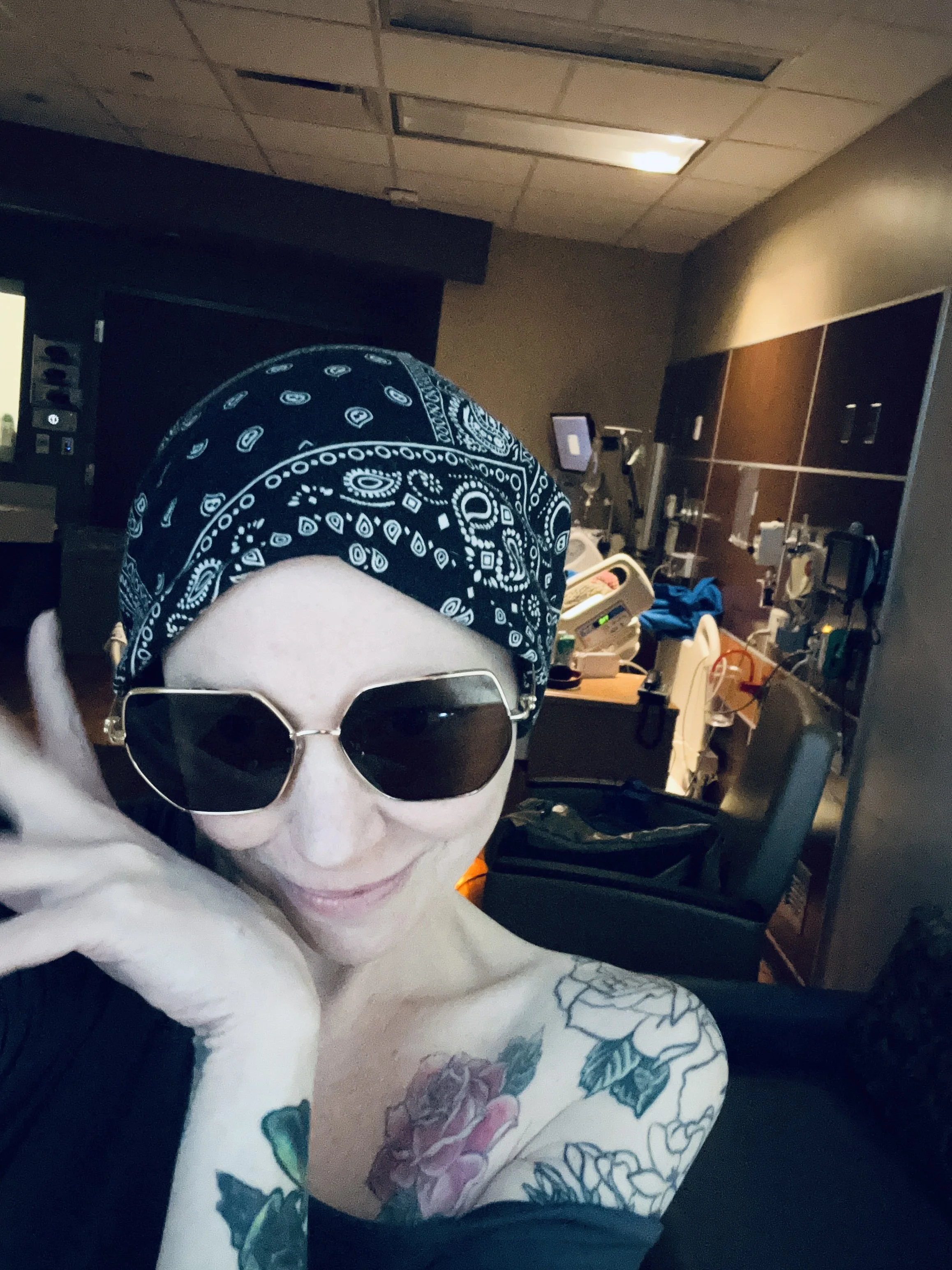 A woman wearing black sunglasses and a black bandana with white patterns is lying in a hospital room. She has tattoos on her shoulder and arm, and is smiling at the camera. The hospital room has medical equipment, monitors, and supplies in the background.