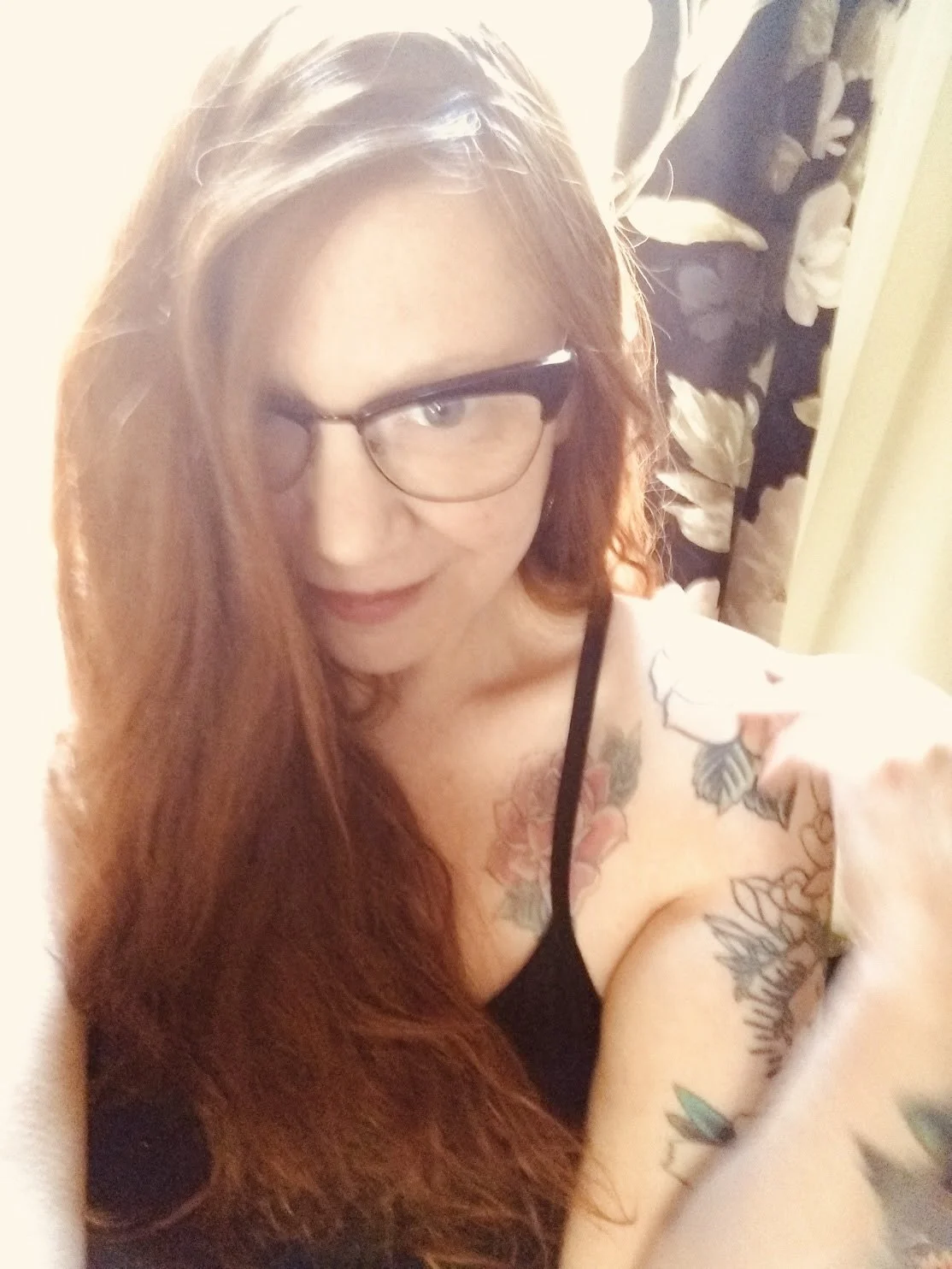 A woman with long red hair and glasses taking a selfie in a room with floral curtains.