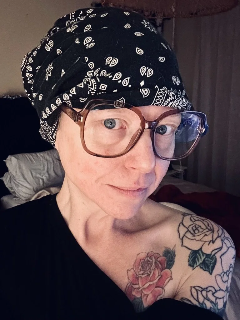 A woman with short blonde hair, wearing a black bandana with a white paisley pattern, large brown glasses, and a black top, smiling. She has colorful floral tattoos on her shoulder.