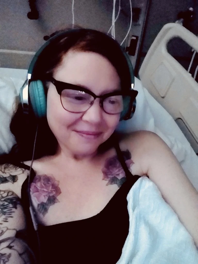 A woman with glasses and dark hair lying in a hospital bed, wearing headphones, a black tank top, and displaying colorful tattoos on her shoulders.