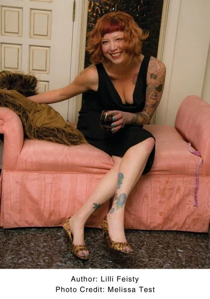 A woman with short red hair and tattoos, wearing a black dress, sitting on a pink sofa in a room with white walls and a door, holding a glass of red wine, smiling.