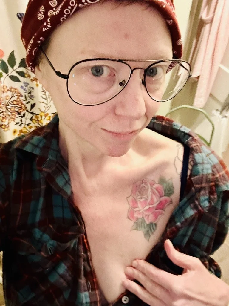 A woman with short hair wearing glasses, a red bandana, and a plaid shirt partially unbuttoned, revealing a tattoo of a pink rose with green leaves on her chest.