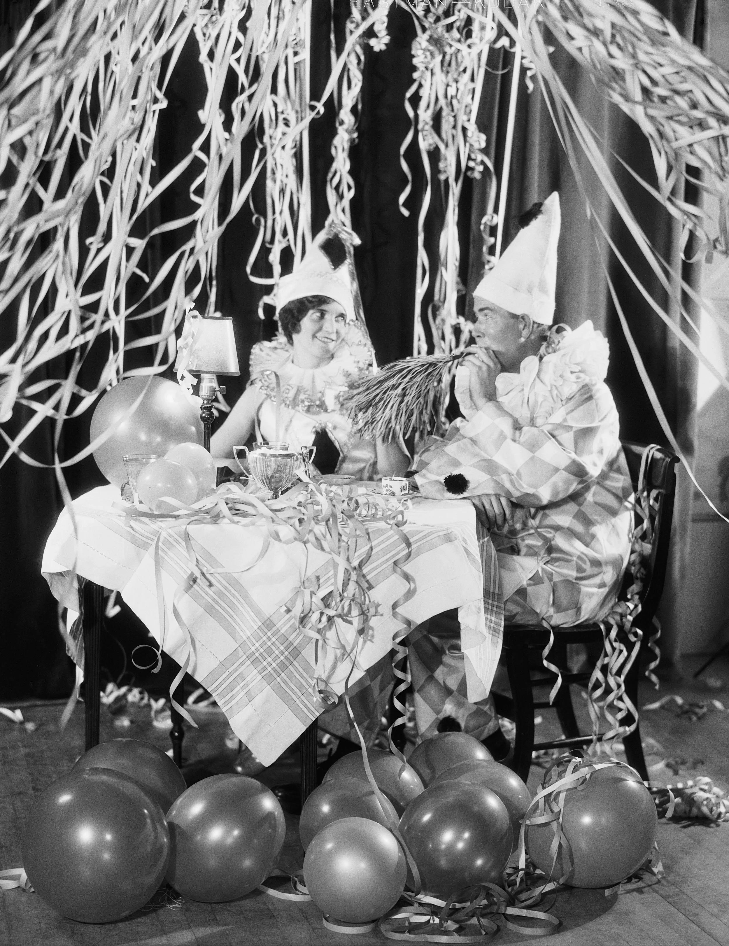Black and white photo of two people dressed as jesters or clowns sitting at a decorated table with balloons, streamers, and paper decorations, with curtains in the background.