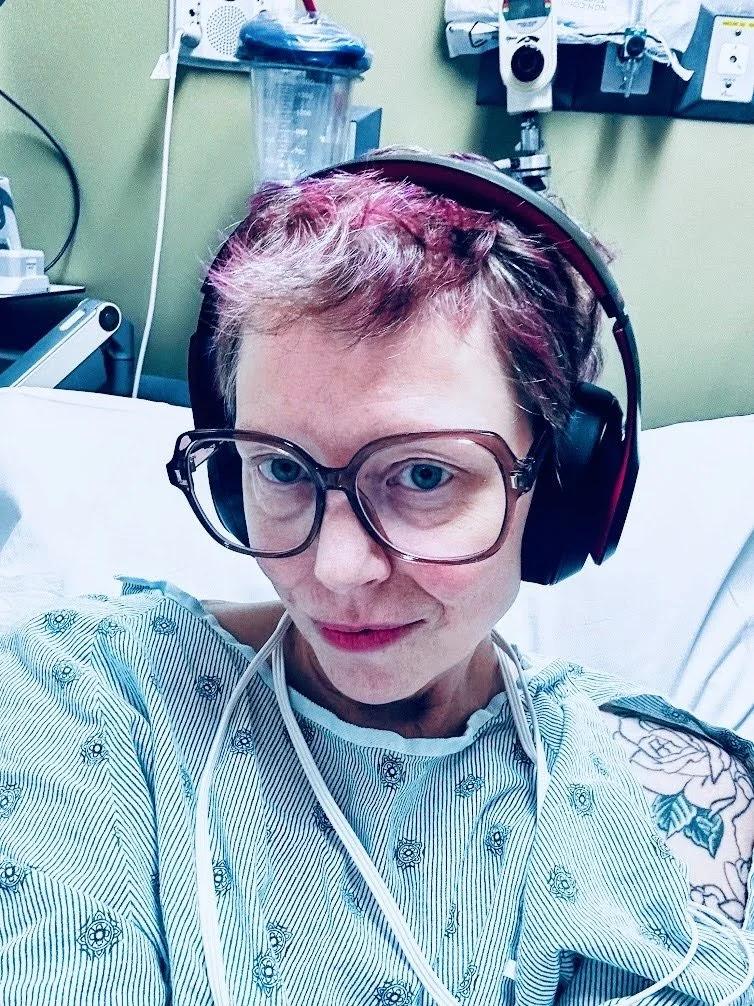Person in hospital gown lying on a hospital bed, wearing large glasses and a headset, with medical equipment and a green wall in the background.