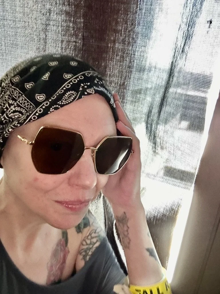 Woman wearing large hexagonal sunglasses, bandana, and a gray shirt, with tattoos on her arms, posing indoors near a textured curtain or wall.