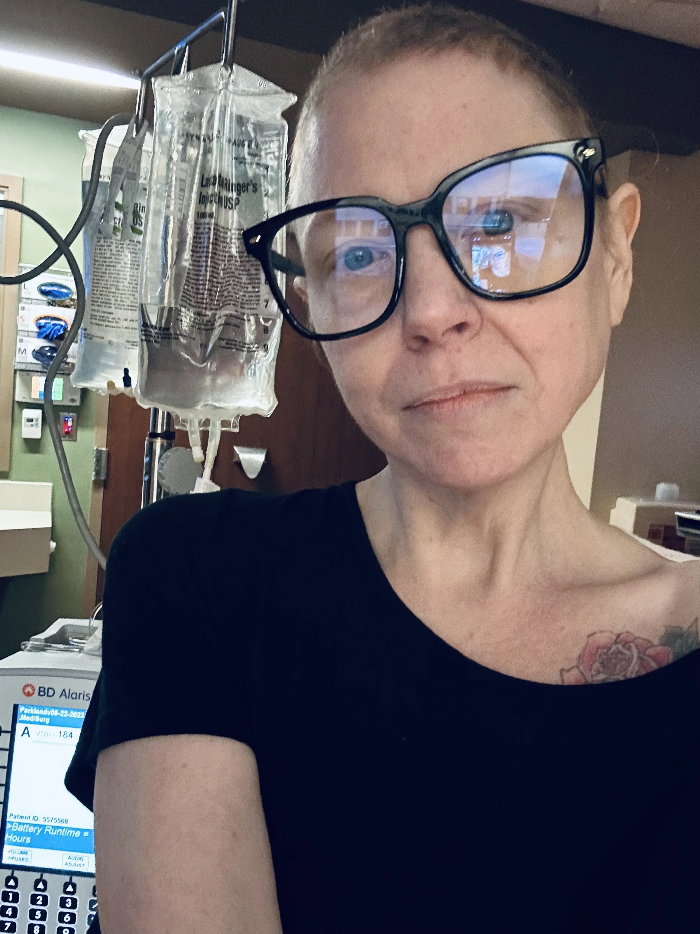 A close-up of a woman in a hospital or medical setting, wearing glasses and a black shirt, with an IV drip bag hanging nearby, and a medical monitor visible in the background.