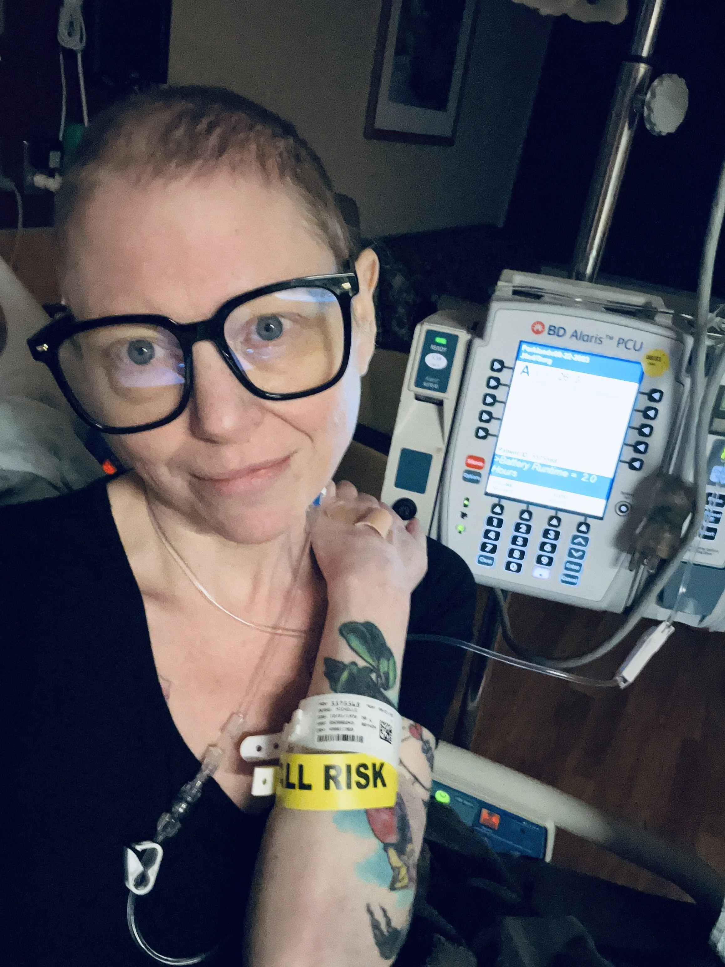 A person with glasses and tattoos in a hospital bed, connected to medical equipment, holding a wristband that reads 'FALL RISK,' with an EKG monitor and medical devices nearby.