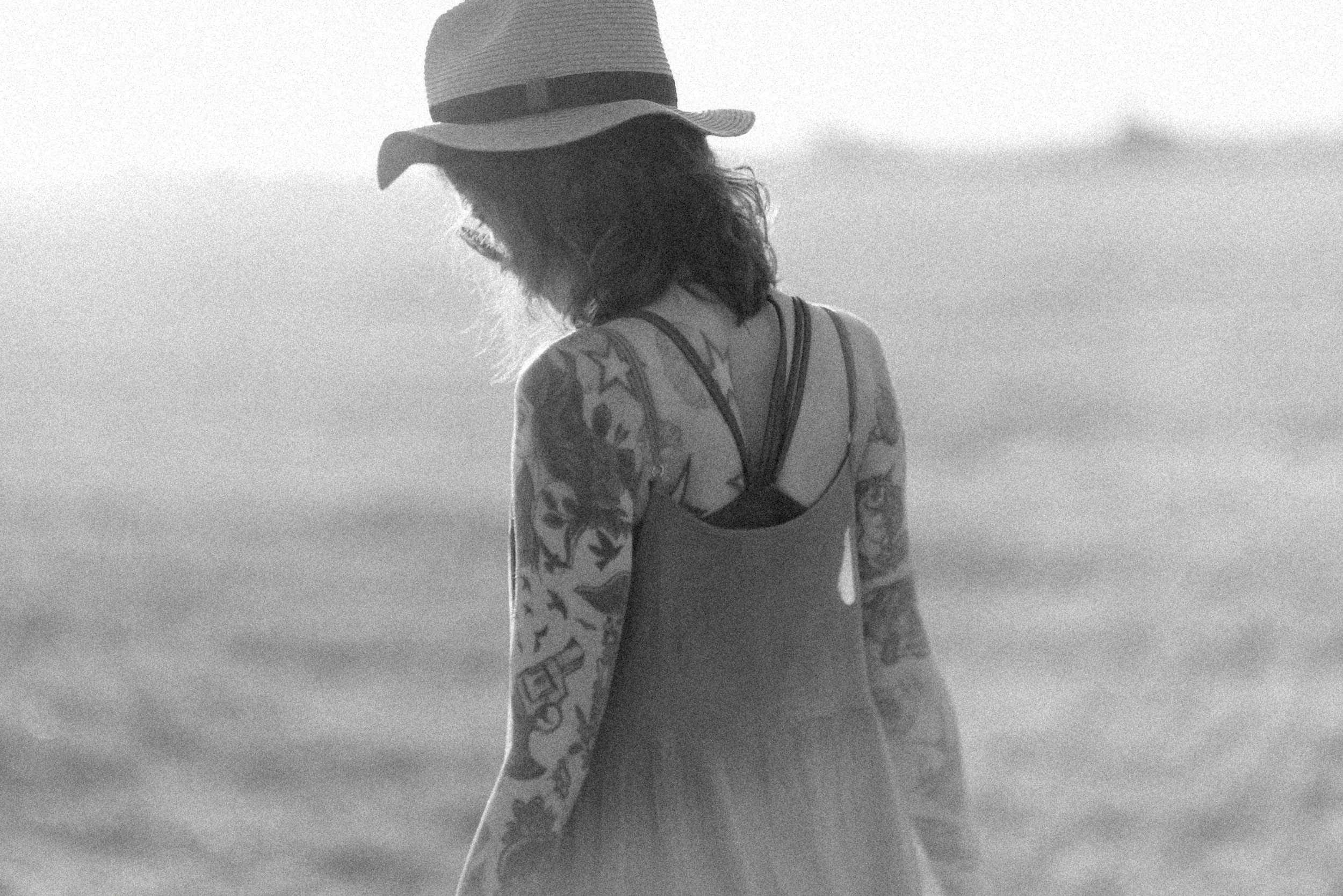 A woman standing on the beach, wearing a wide-brimmed hat, a tattooed sleeve, a tank top, and a patterned kimono, looking down with her back to the camera.