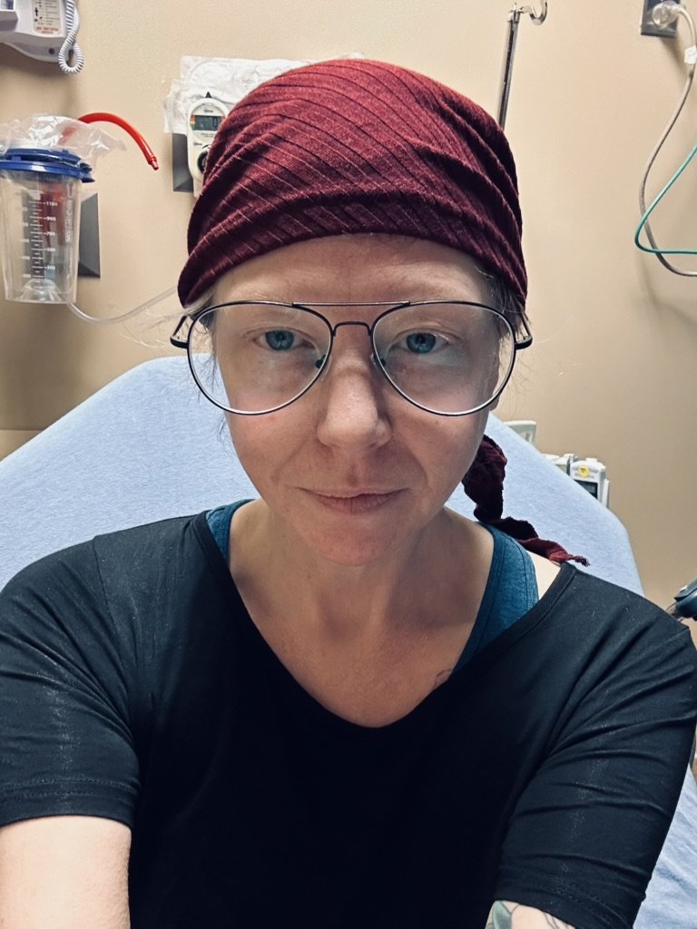 Person wearing glasses and a maroon headscarf sitting on a hospital bed in a medical room.