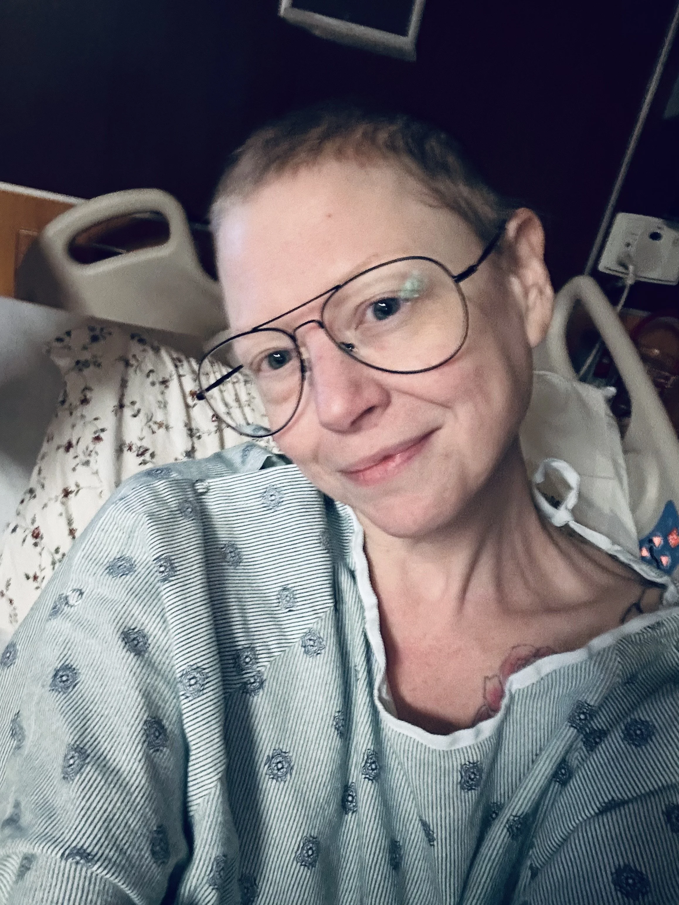 A person lying in a hospital bed with a hospital gown and glasses, smiling at the camera.