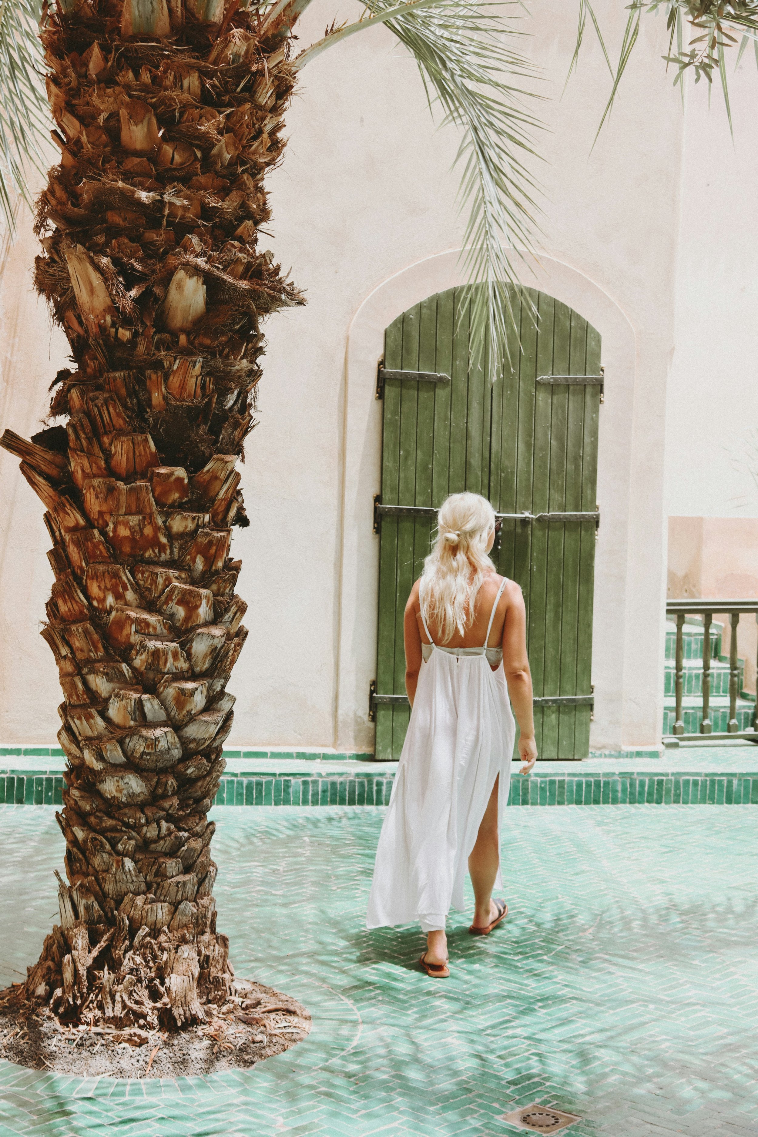 A woman with long blonde hair wearing a white dress and sandals is walking in a courtyard with a green-tiled floor, surrounded by a palm tree, a swimming pool, and a green wooden door.