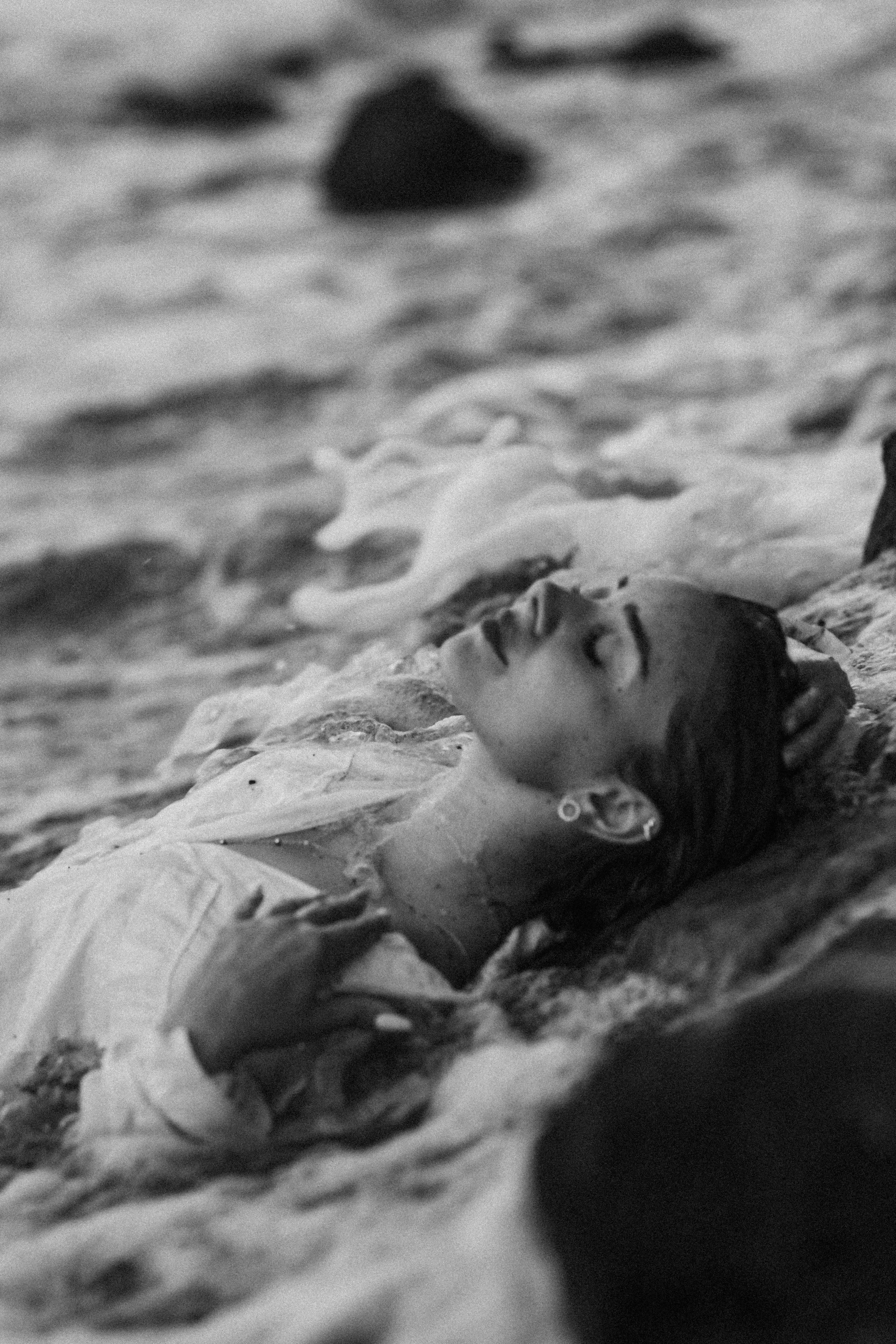A woman lying on the beach in the sand with her eyes closed, wearing earrings and a light-colored top.