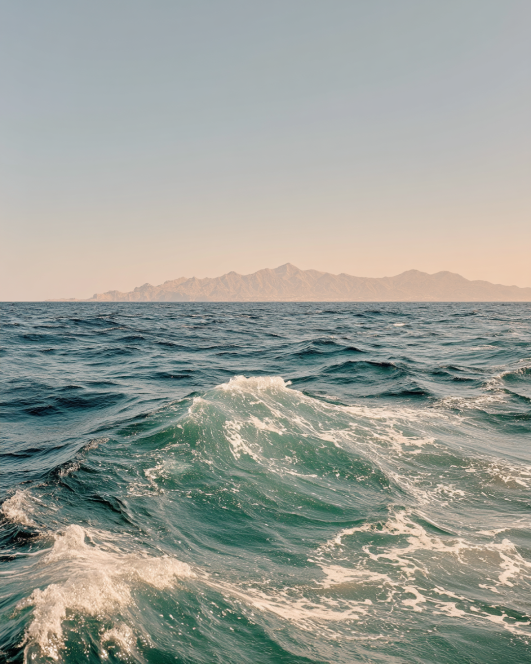 Ocean water with small waves and distant mountains under a clear sky at sunset.