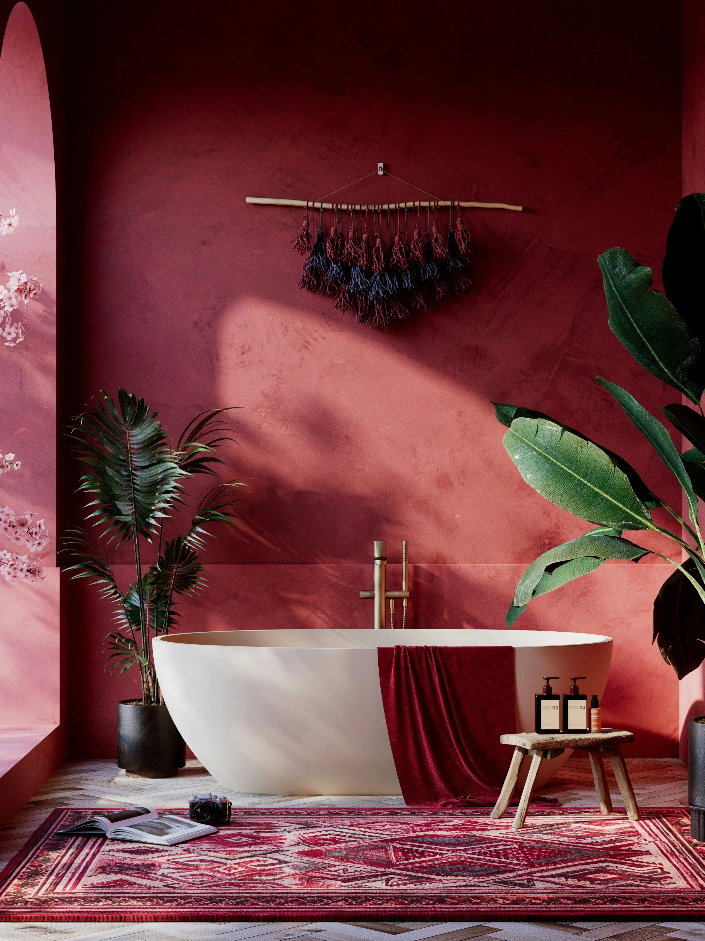 Modern bathroom with a white freestanding bathtub placed on a red and white patterned rug. There are large green houseplants on both sides of the bathtub, and a red towel hangs over the edge. On the right side, a small rustic stool holds black and white toiletry bottles. The deep red wall behind features a textured finish, and above the bathtub, a decorative wall hanging made of a piece of driftwood and tied yarn in black, blue, and red colors. A camera and an open book are on the floor in front of the rug.