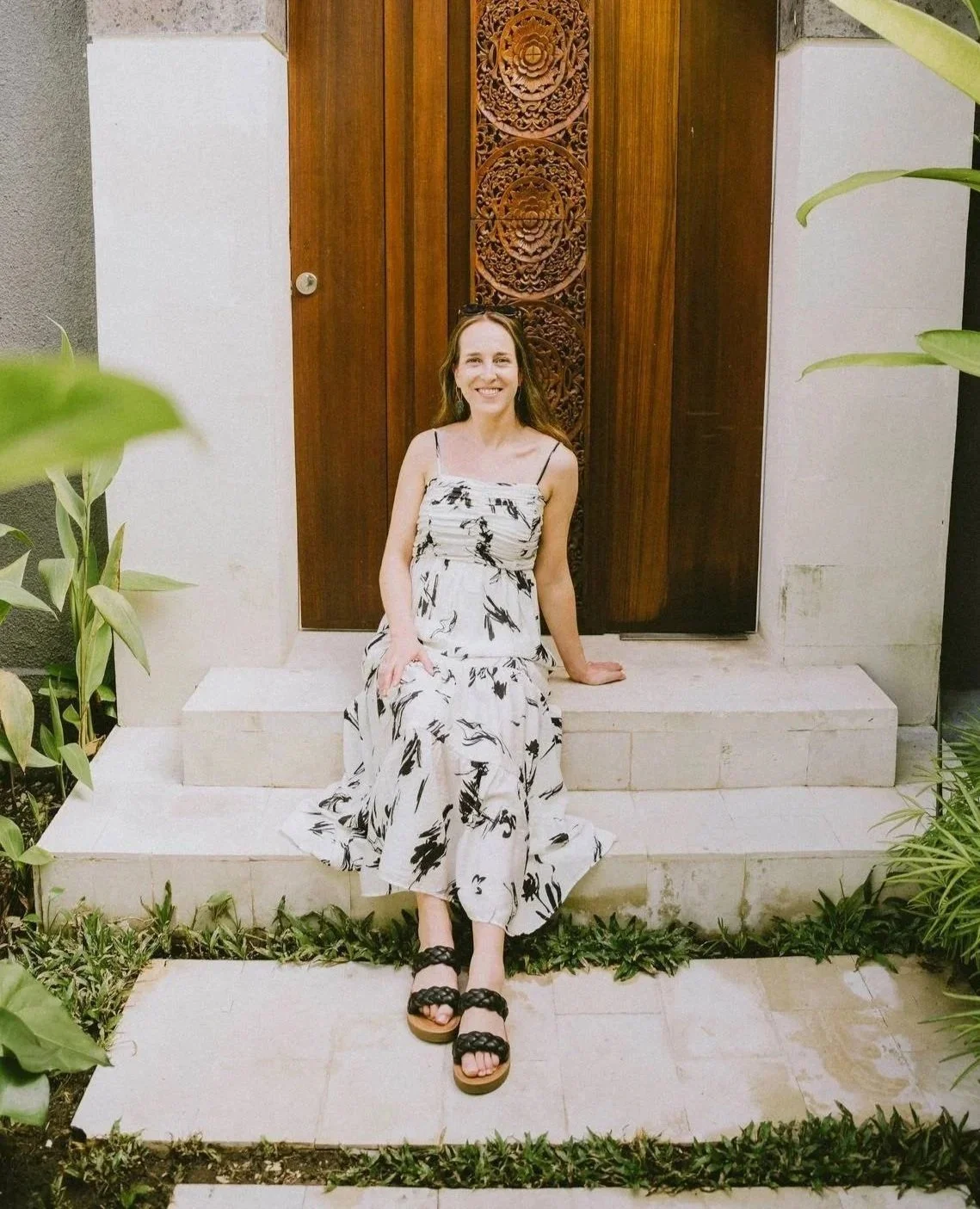 Woman sitting on steps in front of an ornate wooden door, wearing a black and white dress and black sandals, smiling outdoors.