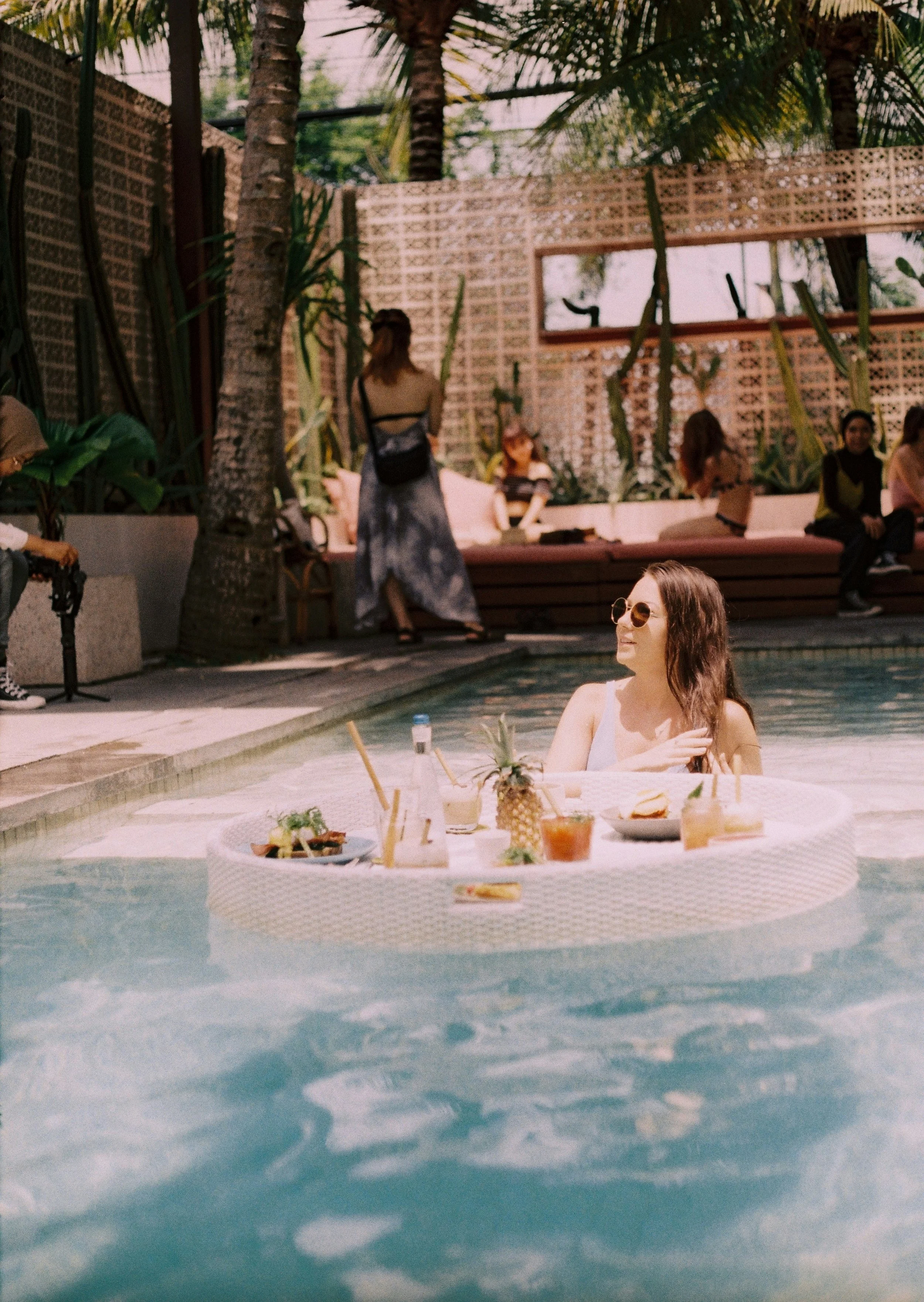 A woman with long brown hair and sunglasses sitting by a poolside table with tropical drinks and a pineapple, in a backyard with a swimming pool, palm trees, and people in the background.