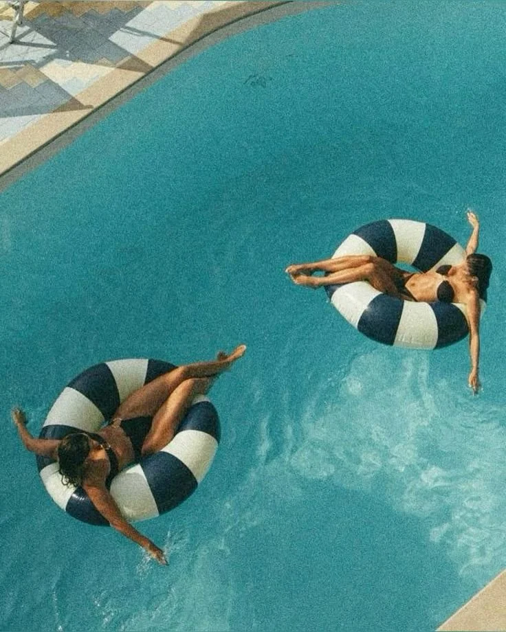 Two women floating on large black and white striped inner tubes in a swimming pool, with one woman in black swimwear and the other in dark swimwear.