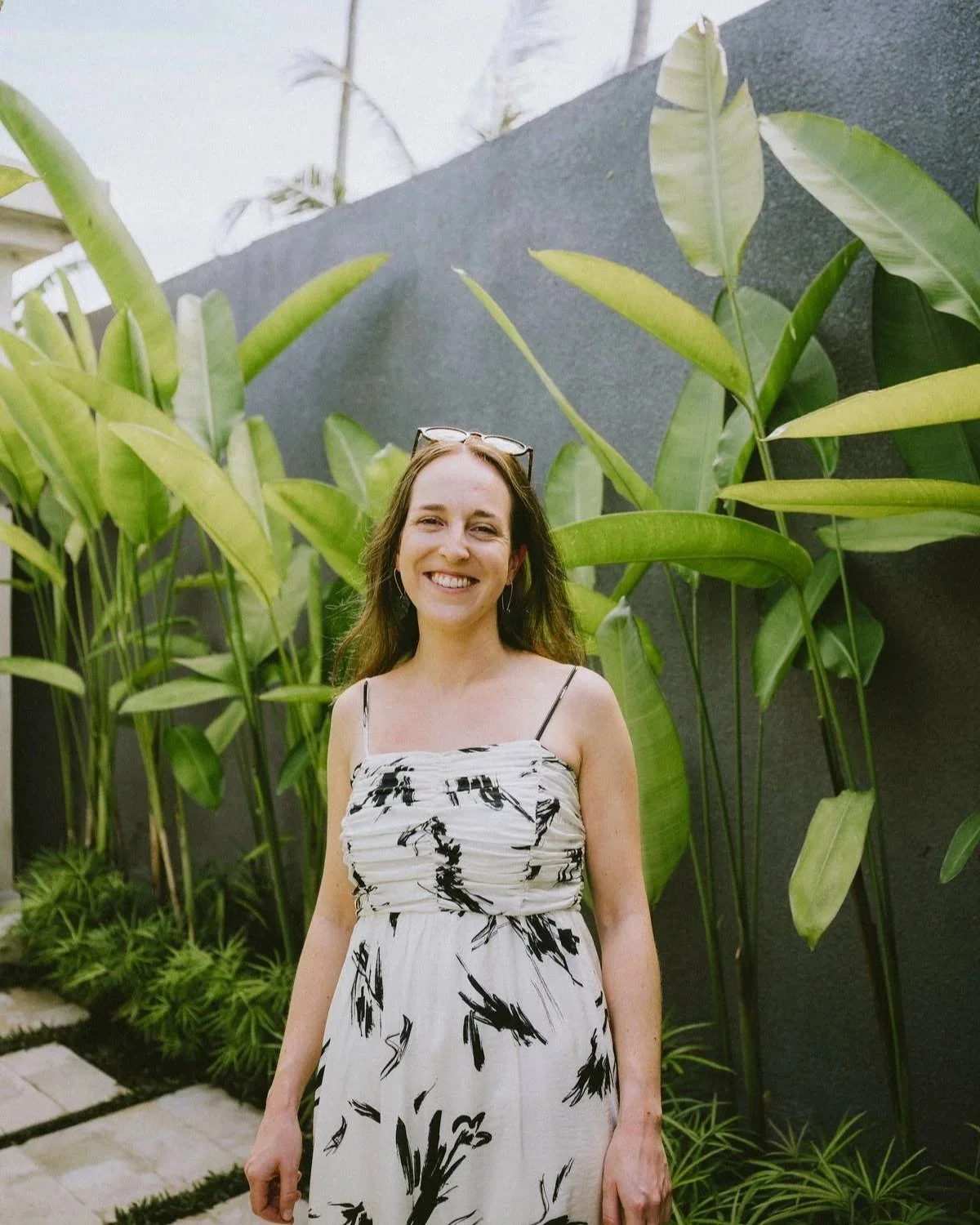 A smiling woman with long brown hair wearing a white dress with black abstract patterns, standing in front of green tropical plants and a dark wall.