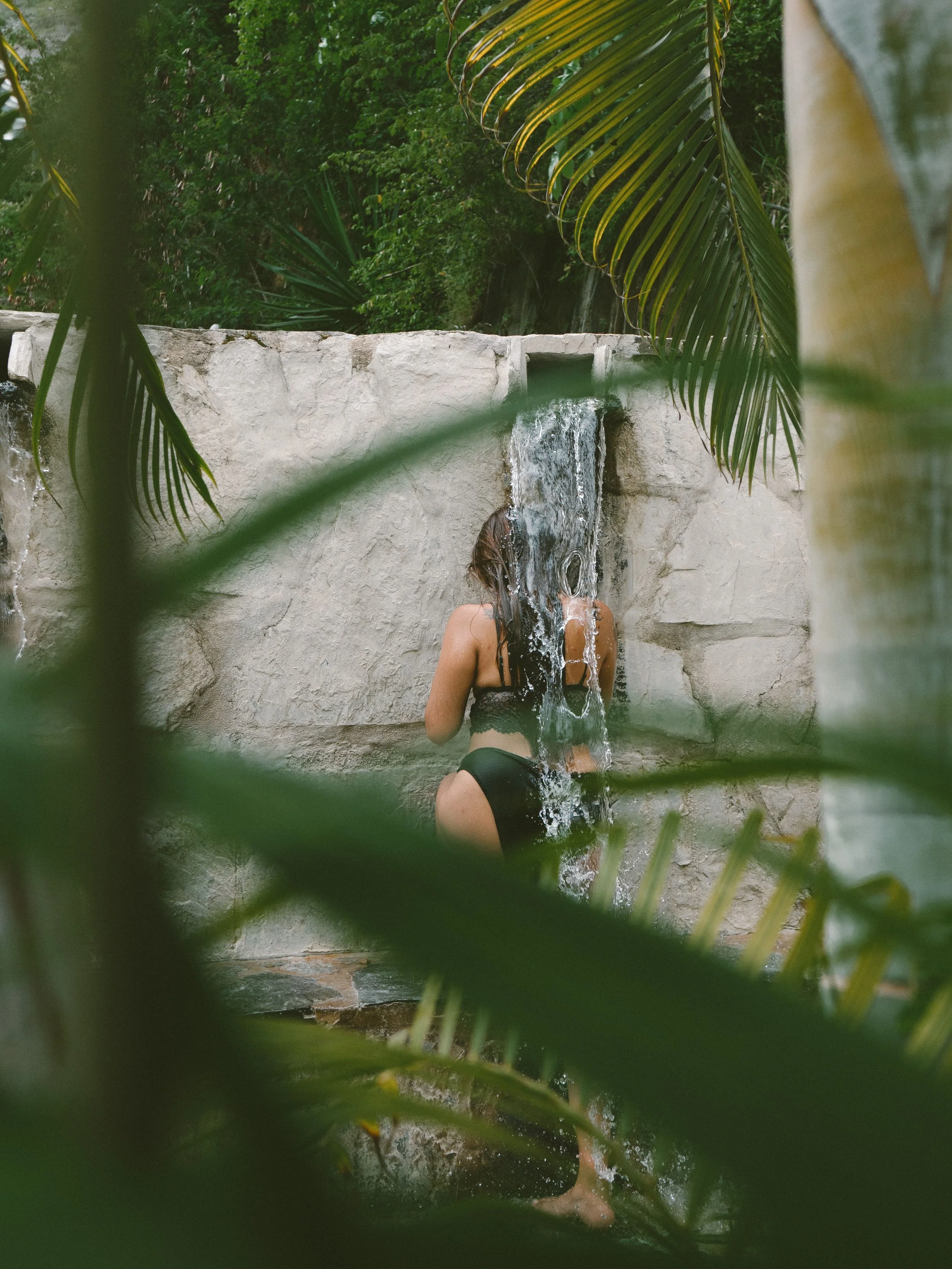 Person with long hair taking a shower outdoors, with water flowing on their head and back, surrounded by green tropical plants.