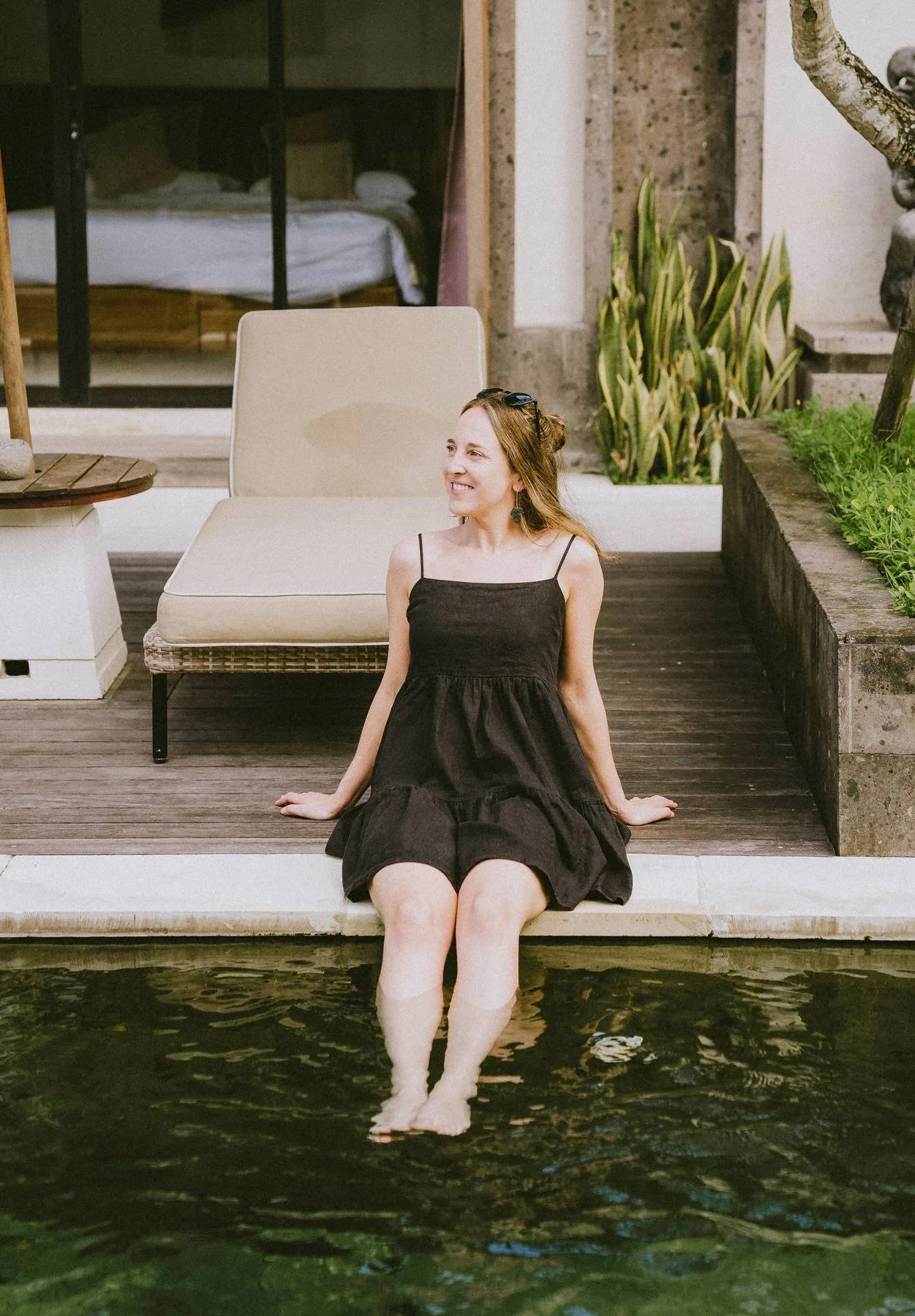 A woman in a black dress sitting on a dock with her feet dipped in a pool of water, smiling and looking to her left. There are green plants and outdoor furniture in the background.