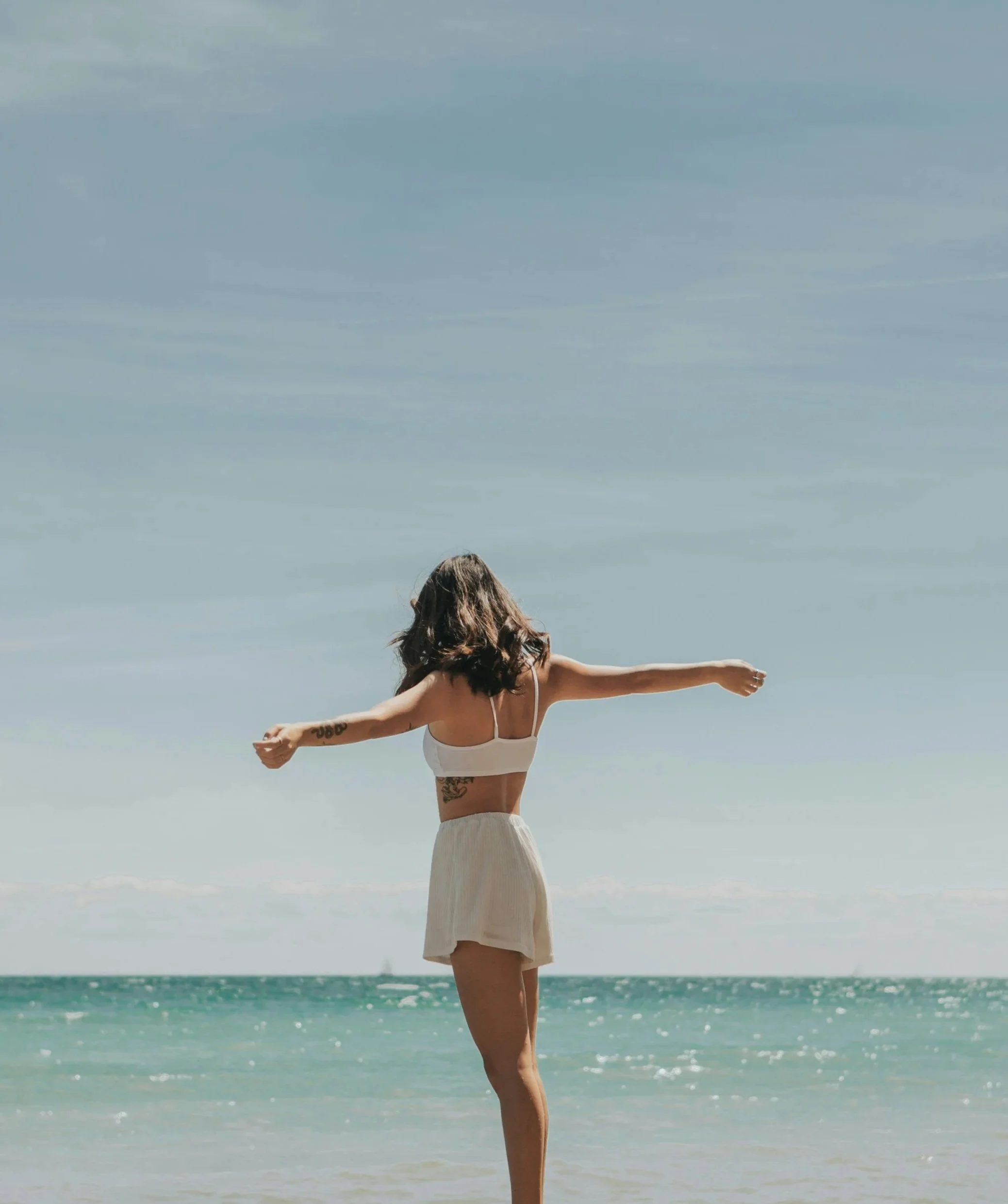 A woman with brown hair in a white top and beige skirt standing on the beach with arms outstretched, facing the ocean and sky.