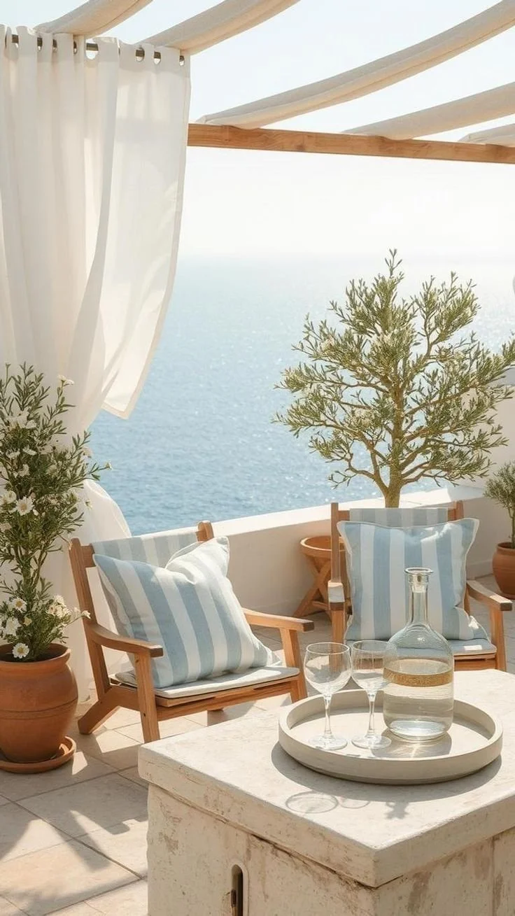 Outdoor balcony overlooking the ocean, decorated with potted plants, striped cushions, and a table with a glass pitcher and two glasses.