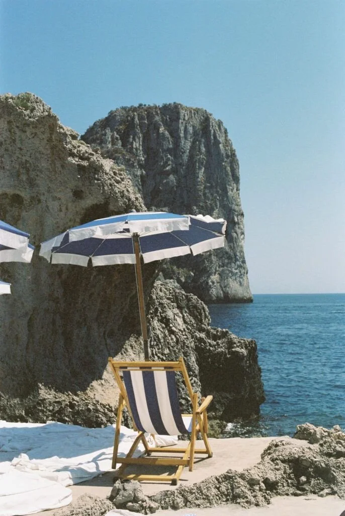 A striped beach chair under an umbrella on a rocky beach with cliffs and blue ocean in the background.
