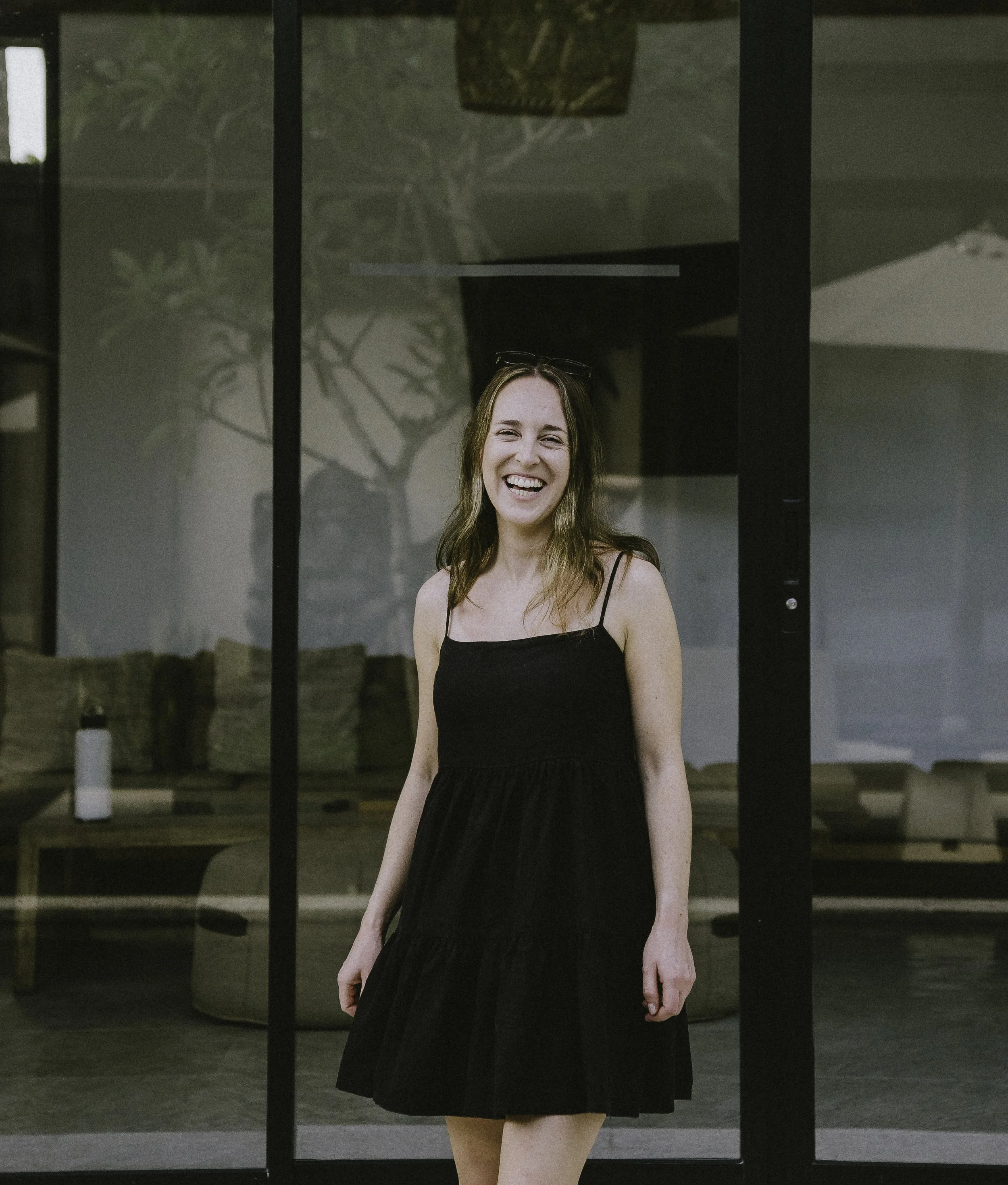 A smiling woman with long light brown hair wearing a black dress, standing outside in front of a modern house with a glass entrance and wooden accents. She appears joyful and relaxed.