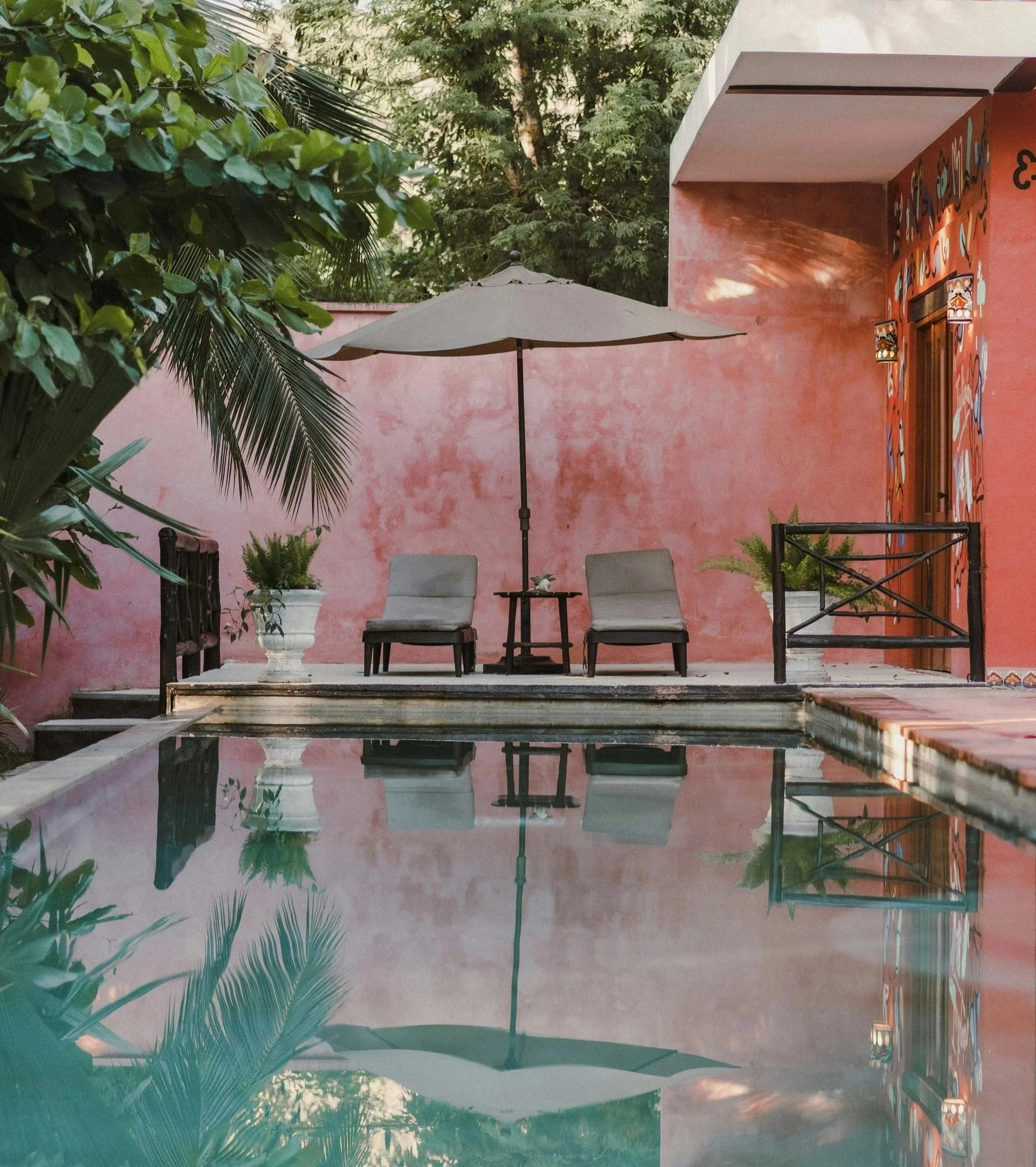 A patio scene with a pink stucco wall, an outdoor table with two chairs, a large patio umbrella, potted plants, and lush green trees in the background, with reflections visible in the pool water.
