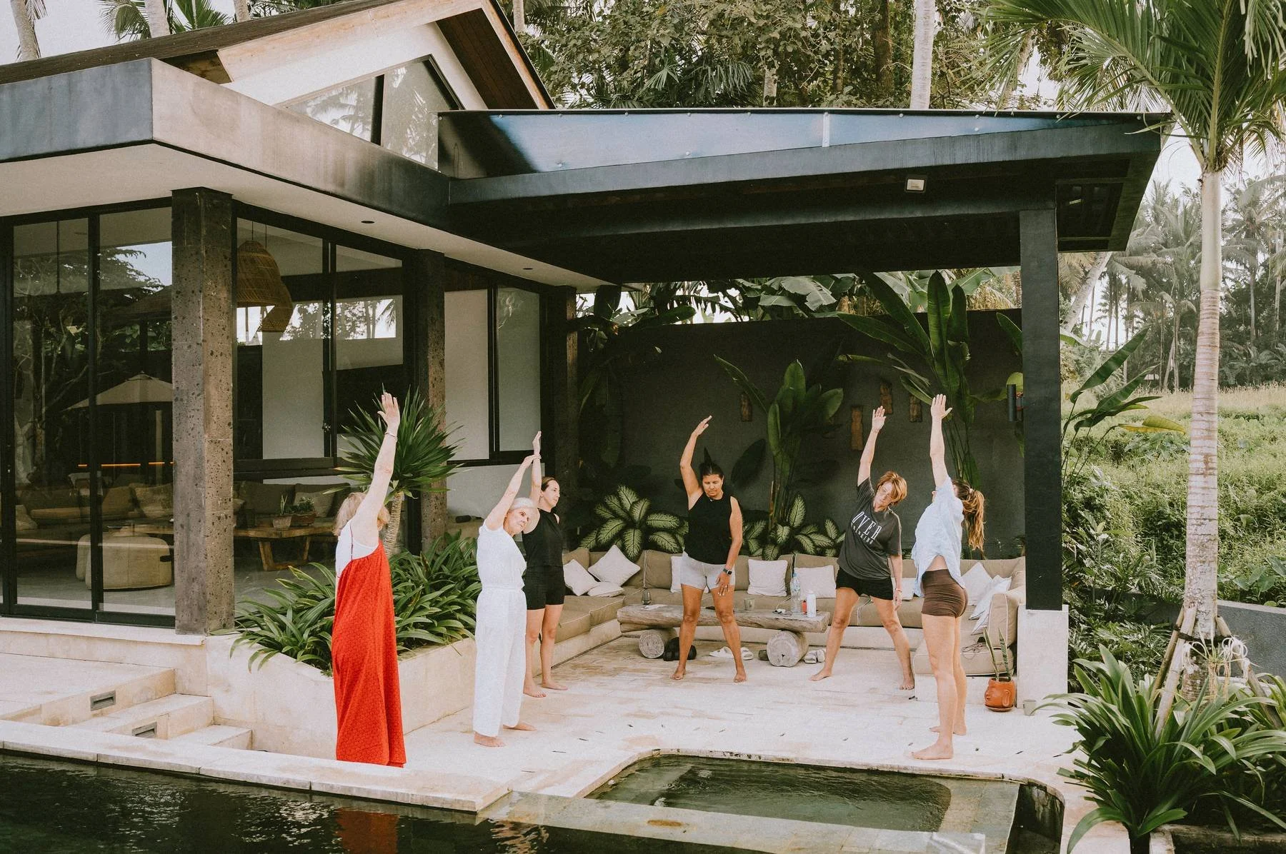 Group of six women practicing yoga outdoors on patio in tropical setting, with modern house and lush greenery in background.