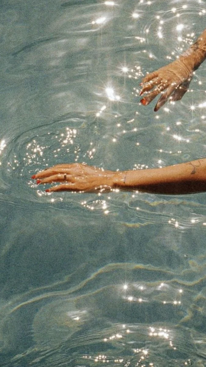 Two hands touching the surface of a sparkling water pool, creating ripples and reflections of light.