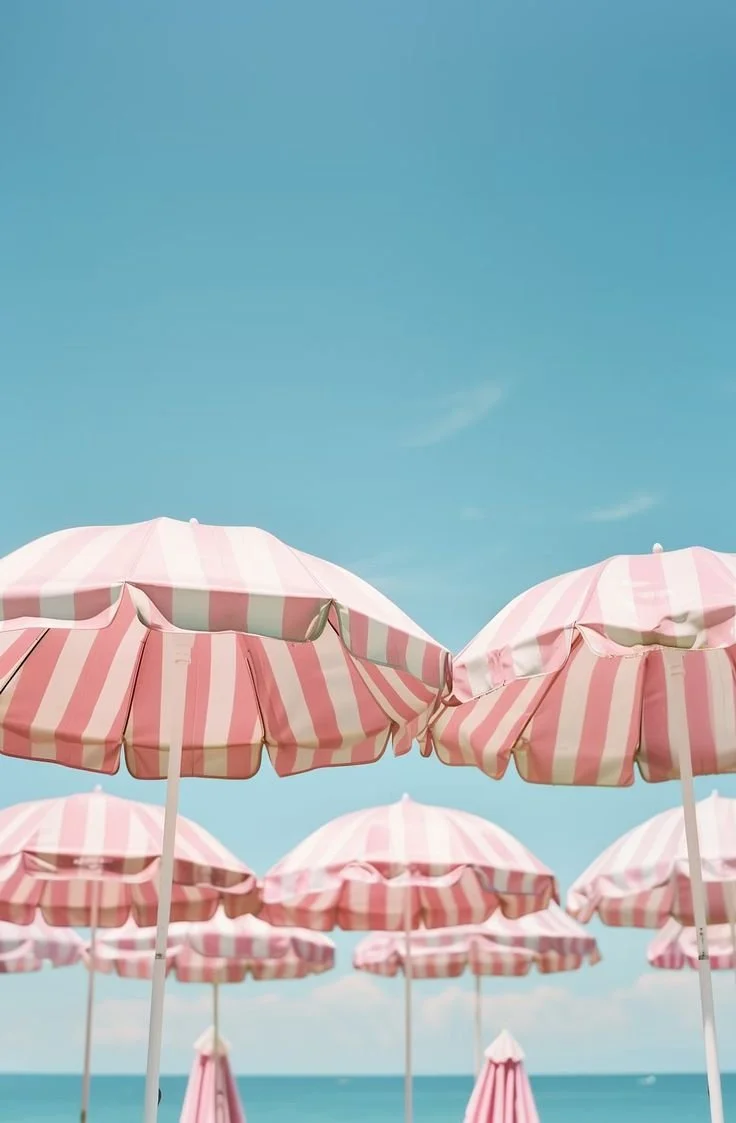 Multiple pink and white striped beach umbrellas against a bright blue sky, with the ocean visible in the background.