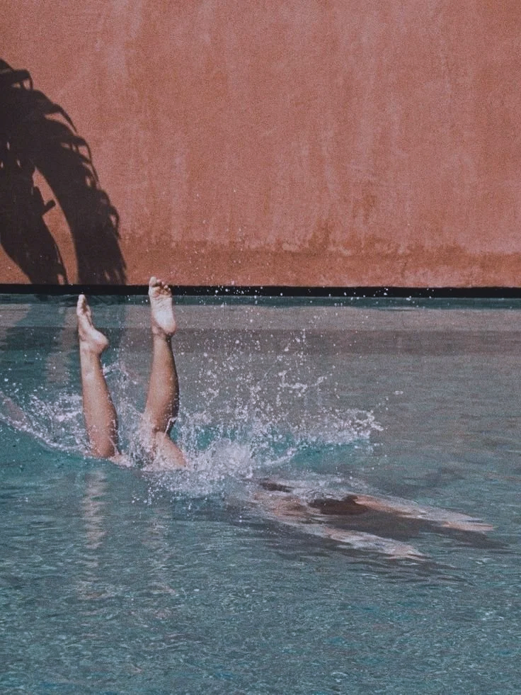 A person in a swimming pool has their legs sticking out of the water, with the rest of their body submerged. A pool wall and a shadow of a plant are visible in the background.