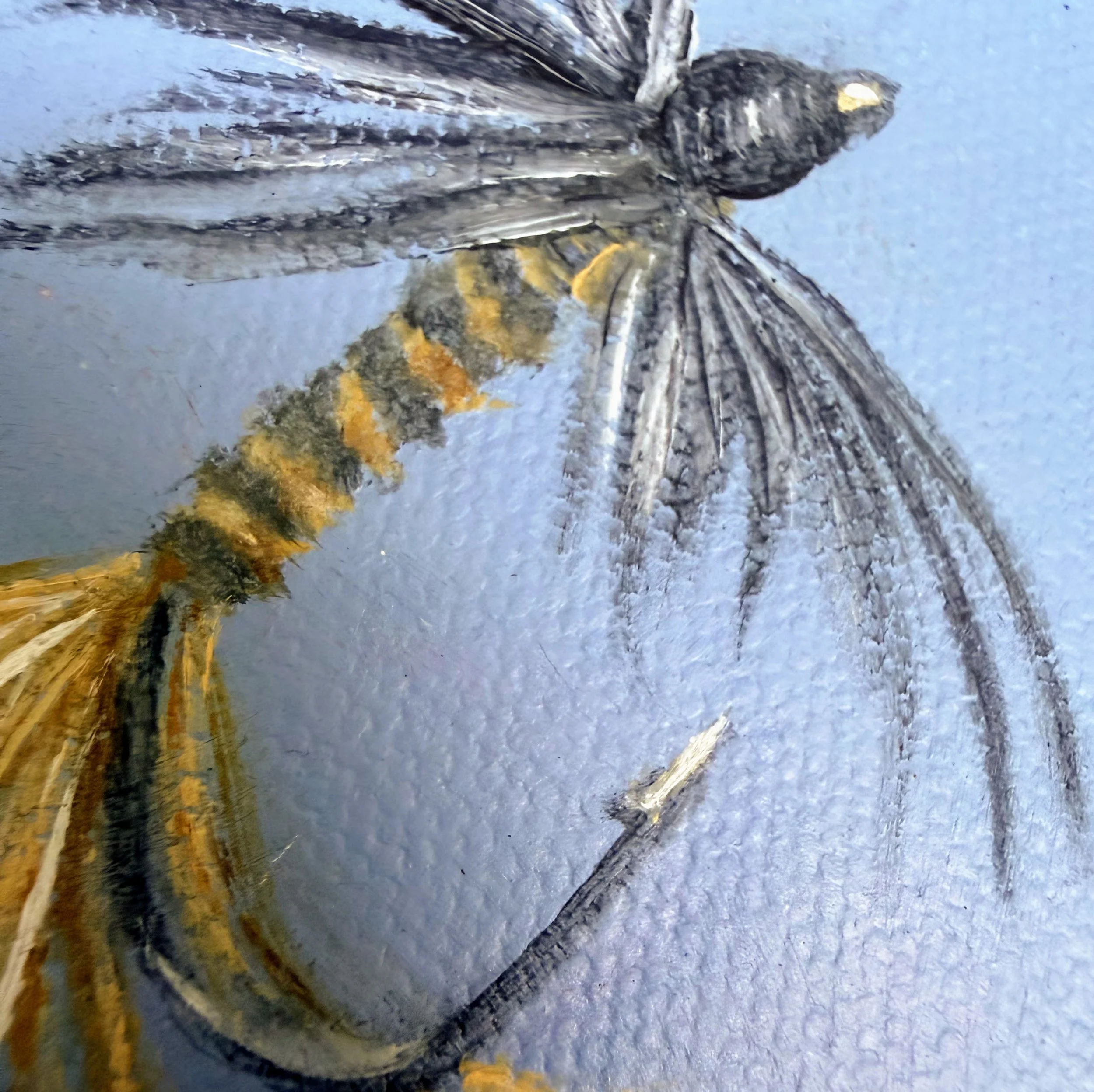 Close-up of a miniature fishing fly painted in oil with fine black feather-like brushwork, striped body detail, pale tail fibers, hook detail, and visible canvas texture.