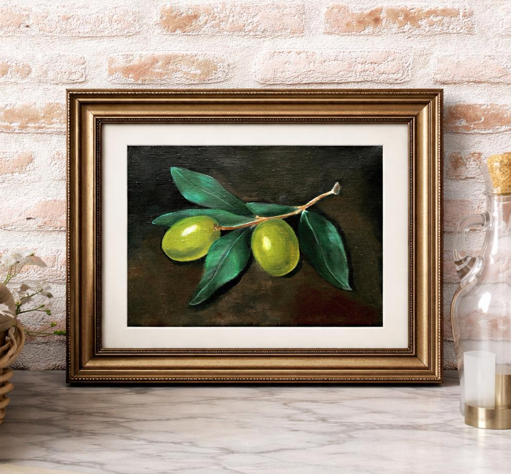 A framed painting of green olives with leaves on a dark background, placed on a wooden surface against a brick wall.